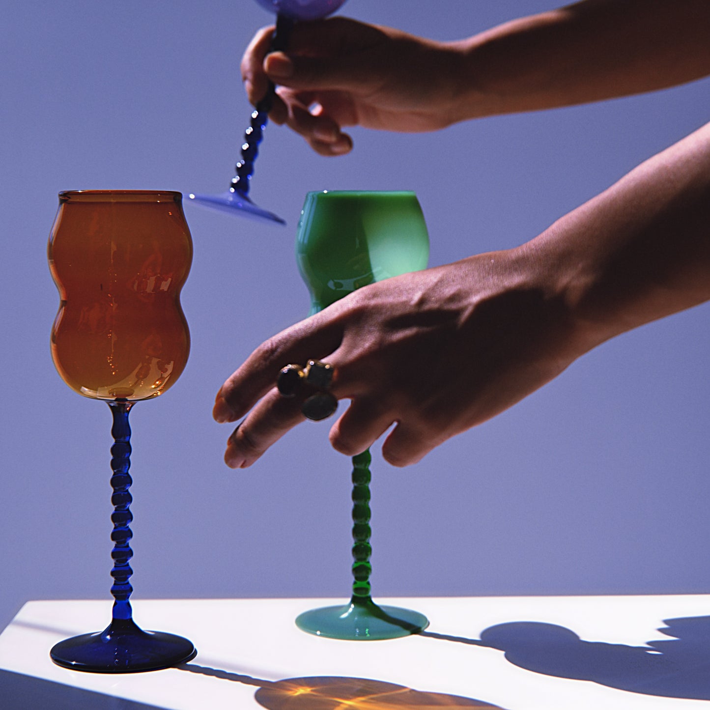 Dreamlike Wine Glass with sculptural design, featuring vibrant orange and green bowls on textured blue stems, held by hands in artistic lighting.